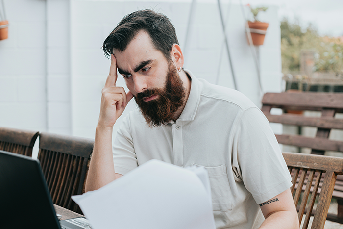 Man with beard looking stressed while reviewing papers and using a laptop, reflecting time for you to get a job dilemma. Man with beard looking stressed while reviewing papers and using a laptop, reflecting time for you to get a job dilemma.
