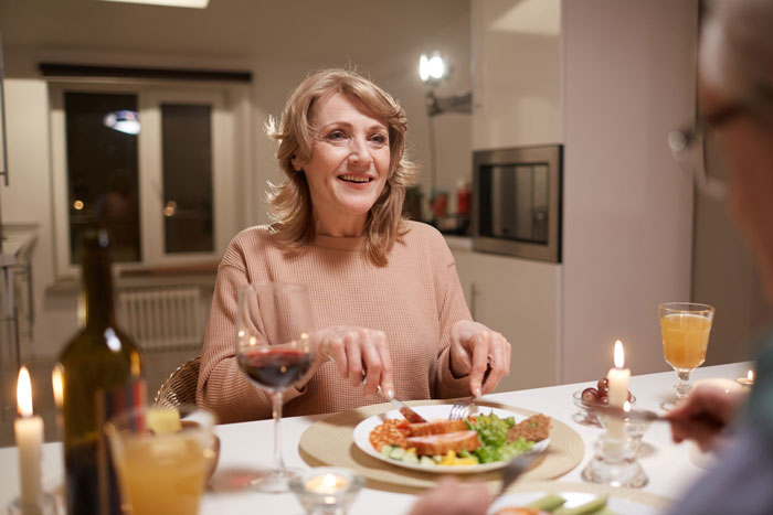 Middle-aged woman sitting at dinner table with food and drinks, representing picky-eater MIL at Thanksgiving dinner. Middle-aged woman sitting at dinner table with food and drinks, representing picky-eater MIL at Thanksgiving dinner.