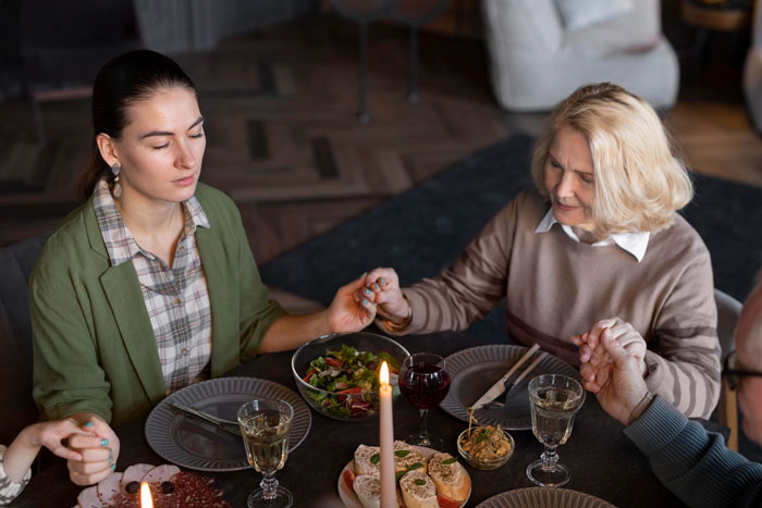 Family holding hands around a Thanksgiving dinner table with wine and salad, highlighting picky-eater MIL food tensions. Family holding hands around a Thanksgiving dinner table with wine and salad, highlighting picky-eater MIL food tensions.