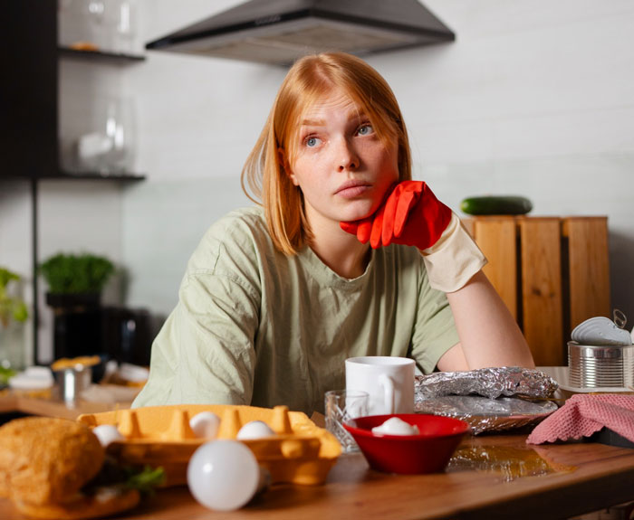 Young woman in kitchen looking frustrated while wearing a red glove, surrounded by food and cooking items reflecting picky-eater MIL issues Young woman in kitchen looking frustrated while wearing a red glove, surrounded by food and cooking items reflecting picky-eater MIL issues