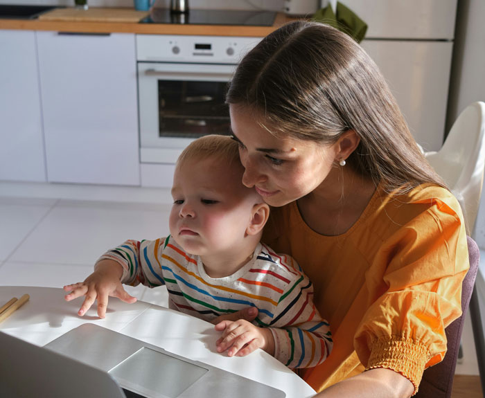 Aunt babysitting a toddler in a kitchen, balancing childcare responsibilities while spending time on a laptop.