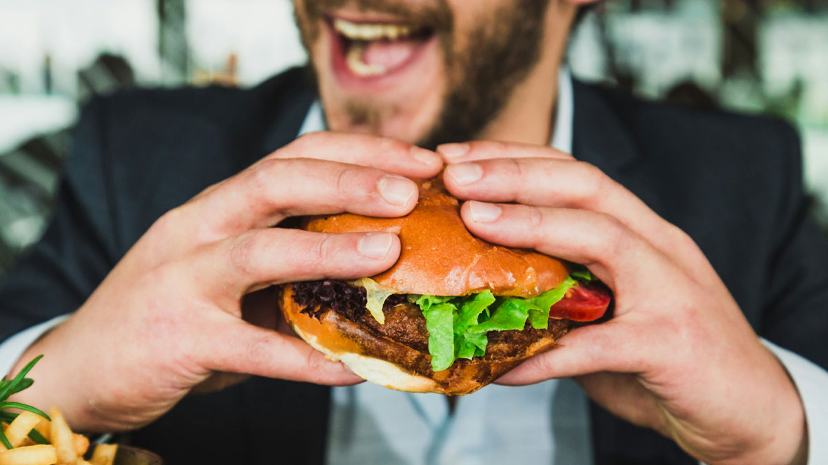 Man in business attire holding a beef burger with lettuce and tomato, smiling before taking a bite.