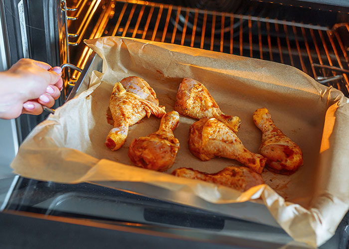 Hand placing a tray of seasoned chicken drumsticks into an oven, highlighting conflicts over strict vegan kitchen rules.