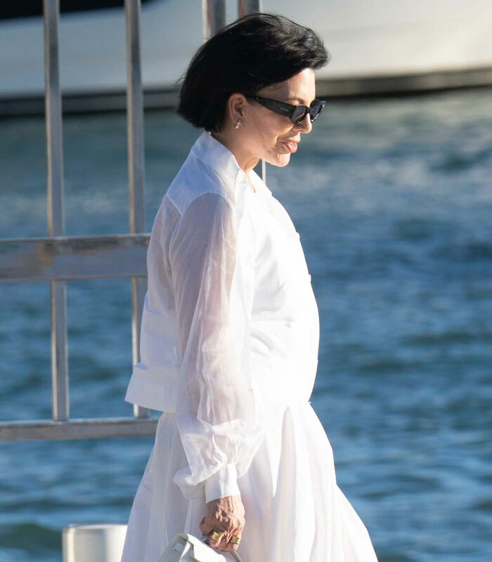 Kris Jenner in white outfit and sunglasses, walking outdoors by water, hands visible after viral $100k facelift news. Kris Jenner in white outfit and sunglasses, walking outdoors by water, hands visible after viral $100k facelift news.