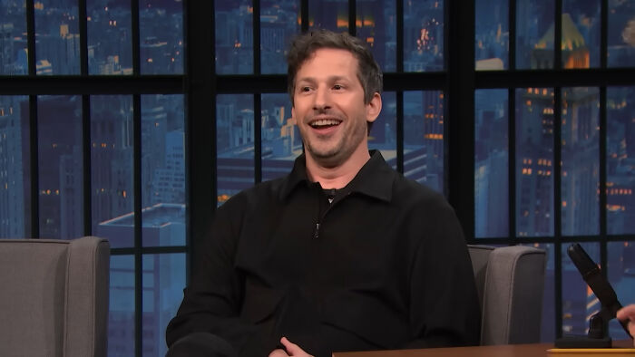 Man smiling and talking on a late night talk show set sharing fascinating secrets about celebrities from insiders.