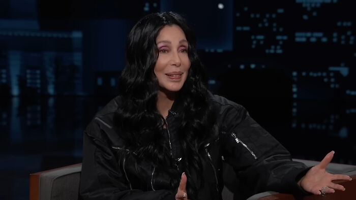 Female celebrity with long black hair in a dark jacket sharing fascinating secrets about celebrities on a talk show.