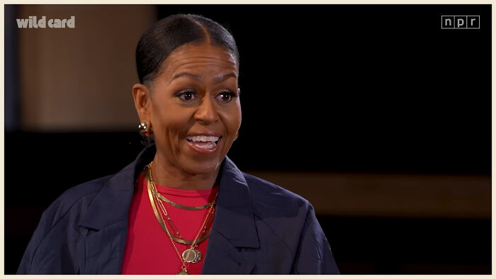 Woman sharing fascinating secrets about celebrities, wearing layered gold necklaces and a dark jacket in an interview setting.