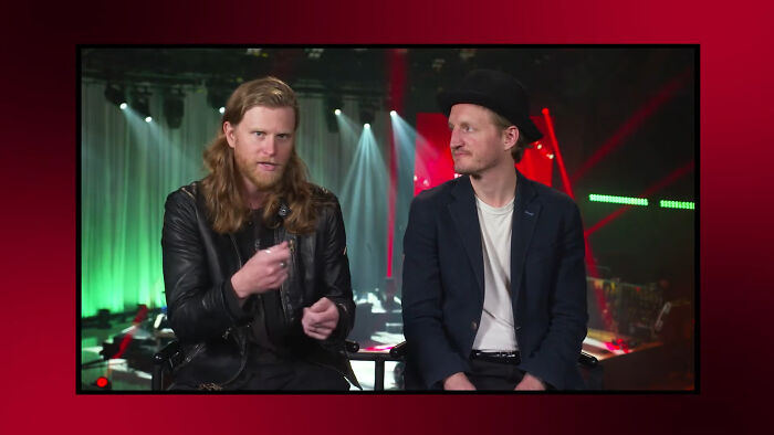 Two men with serious expressions discussing celebrities people think suffer during a live event with stage lights behind them.