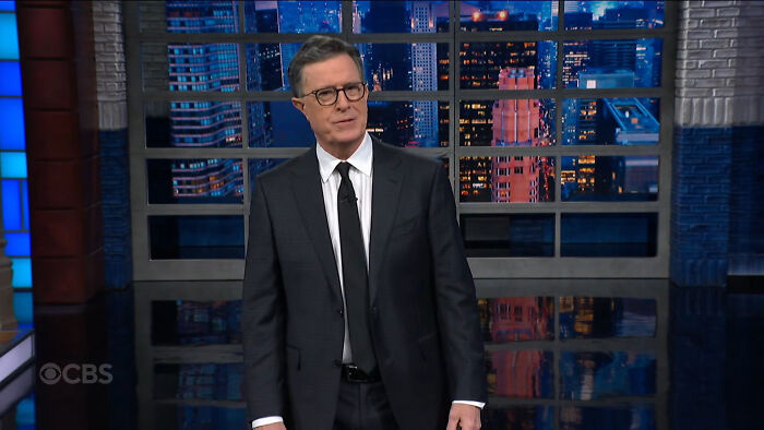 Host in a dark suit and glasses standing on a TV set with cityscape windows, discussing celebrities people think suffer.