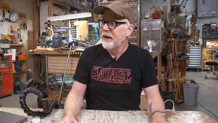 Man wearing glasses and a cap sharing fascinating secrets about celebrities while sitting in a workshop filled with tools.