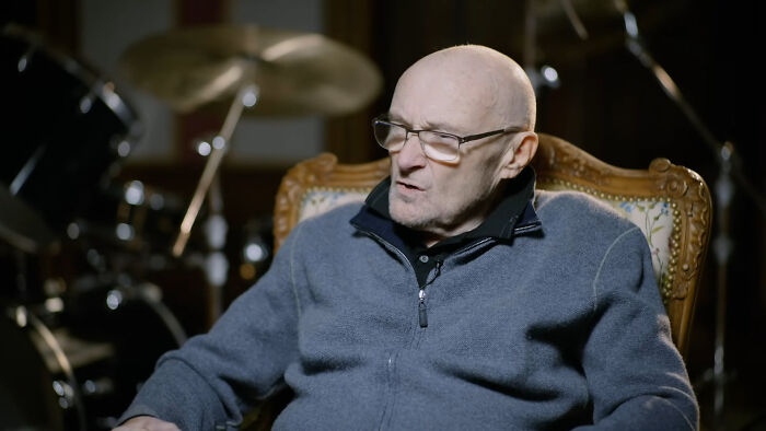 Elderly man wearing glasses and a grey jacket sitting thoughtfully in a vintage chair with a drum set in the background.