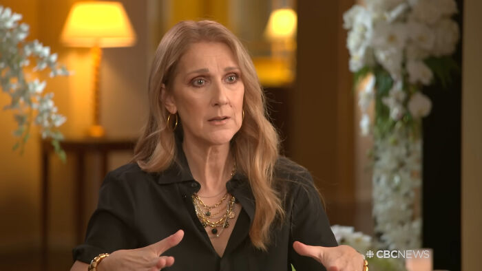 A woman with long blonde hair in a black shirt discussing celebrities people think suffer in a softly lit room.