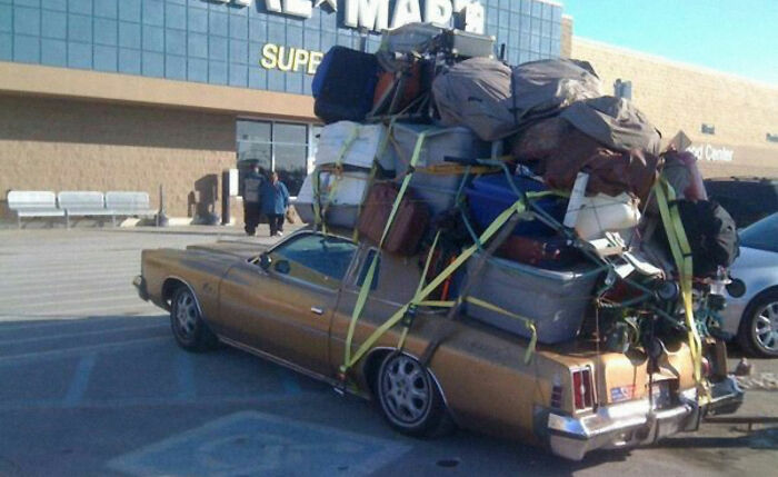 Old car heavily overloaded with numerous bags and suitcases strapped on top in a Walmart parking lot, depicting Walmart people.