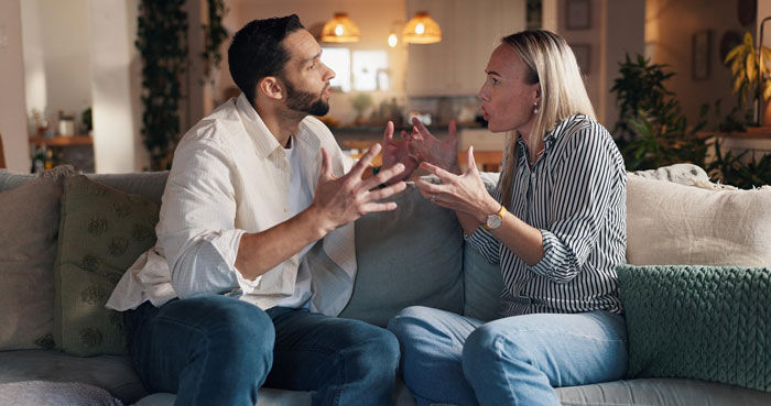 Couple arguing intensely on a couch over debt and clashing values before wedding expenses cause relationship issues.