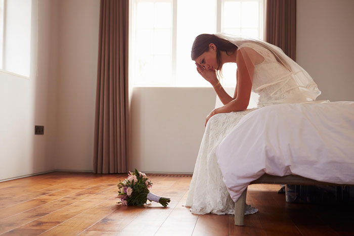 Bride in wedding dress sitting on bed, looking distressed with a bouquet on the wooden floor, reflecting wedding night shock.