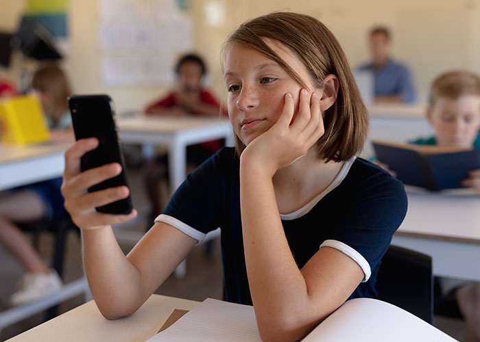 Young girl in classroom looking at smartphone, illustrating unintended consequences of drunken jealousy in everyday life.