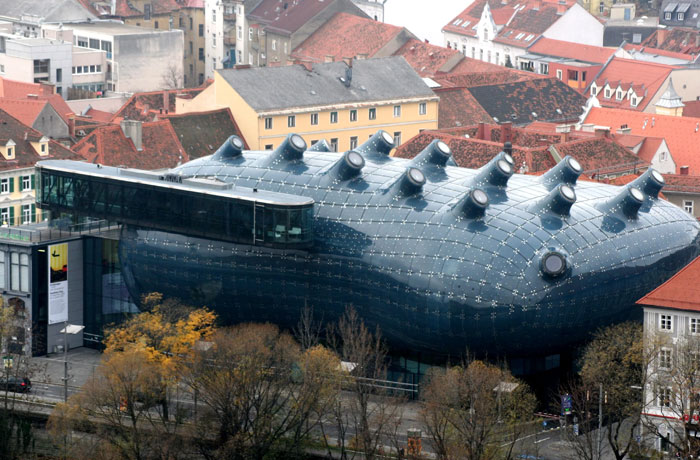 Futuristic bread-shaped building with rounded protrusions, glass extension, surrounded by traditional European rooftops and trees.