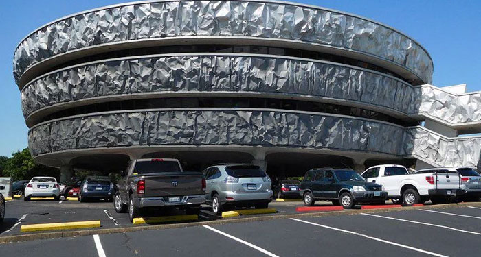 Modern building with circular layered design and crumpled metal facade, located above a busy parking lot with several cars.