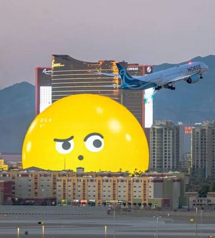 Large yellow emoji-shaped building with a skeptical expression among cityscape as airplane takes off above it.