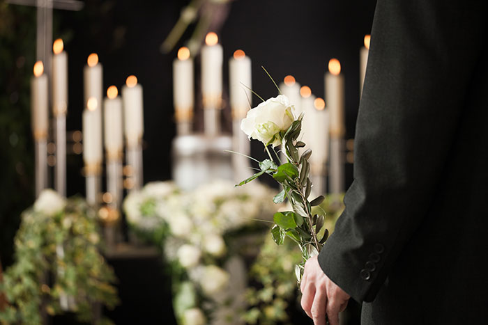 Person in dark suit holding single white rose at a candlelit funeral setting with blurred floral arrangements in background