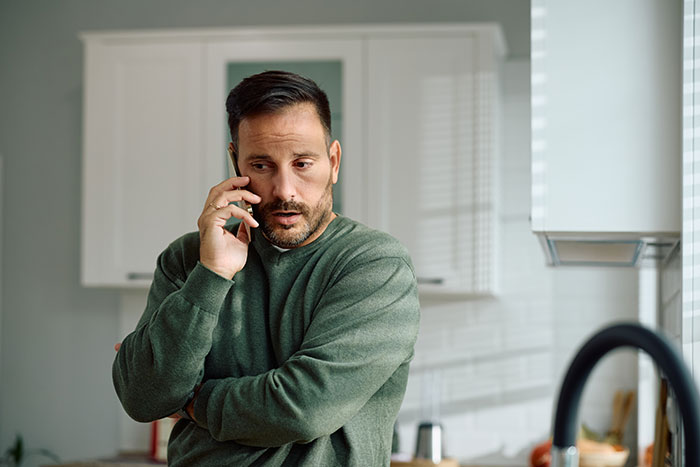 Man in green sweater making a phone call in kitchen, looking concerned while using a strange excuse for missing work