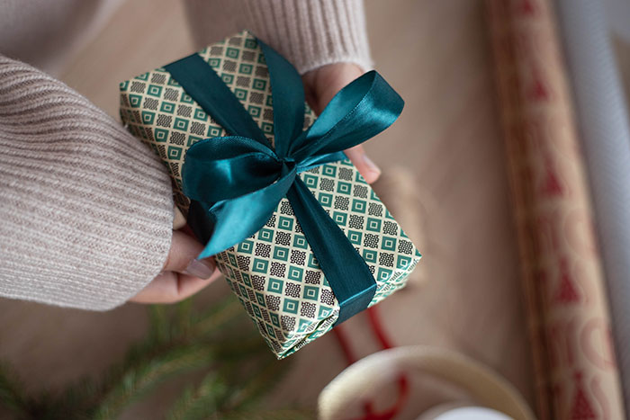 Person holding a neatly wrapped white elephant gift with a dark green ribbon in a cozy indoor setting.