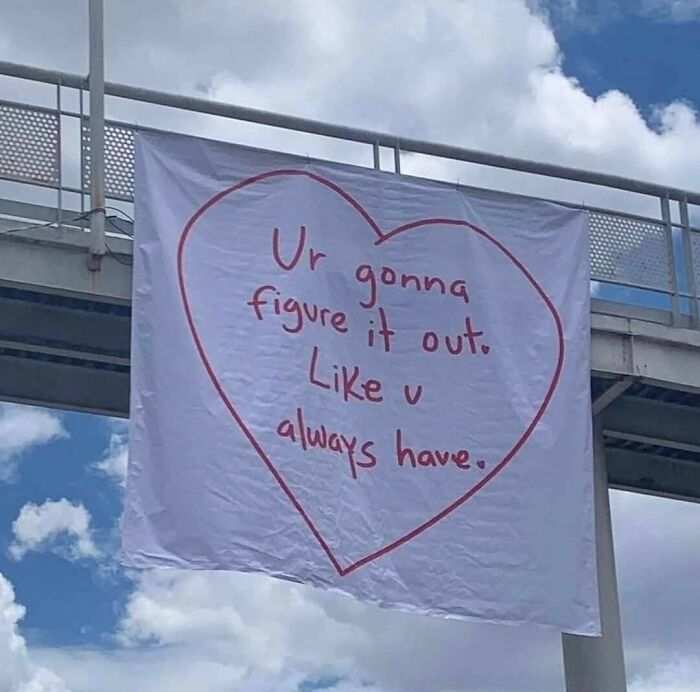 Heart-shaped sign with encouraging message hanging outdoors against a blue sky in a sweet and wholesome pics style.
