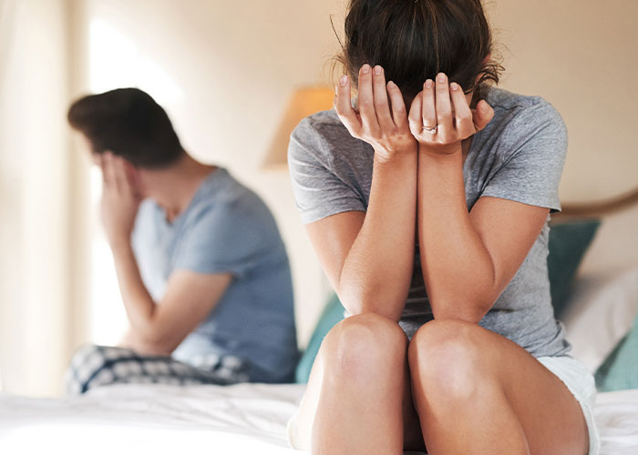 Wife of 10 years covering her face in distress while husband sits on bed, both appearing emotionally upset after separation.