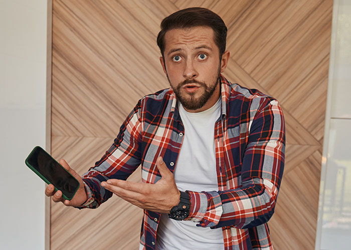 Man in plaid shirt holding phone and looking shocked, illustrating a wife ruining husband’s reputation after leaving him. Man in plaid shirt holding phone and looking shocked, illustrating a wife ruining husband’s reputation after leaving him.
