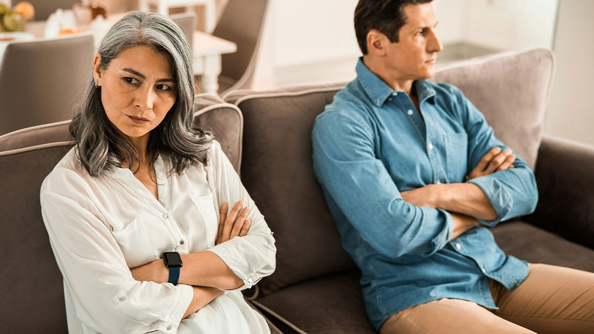 Middle-aged dad and wife sitting apart on a couch with crossed arms, showing tension after a relationship conflict.