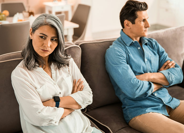 Middle-aged couple sitting apart on couch with arms crossed, depicting dad calling out wife over daughter treating boyfriend like ATM.