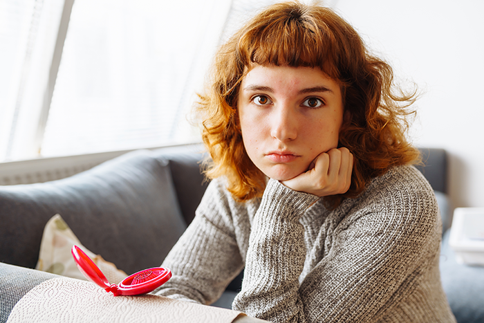 Young person with red hair and gray sweater looking serious, reflecting on family struggles after coming out and facing transphobia.