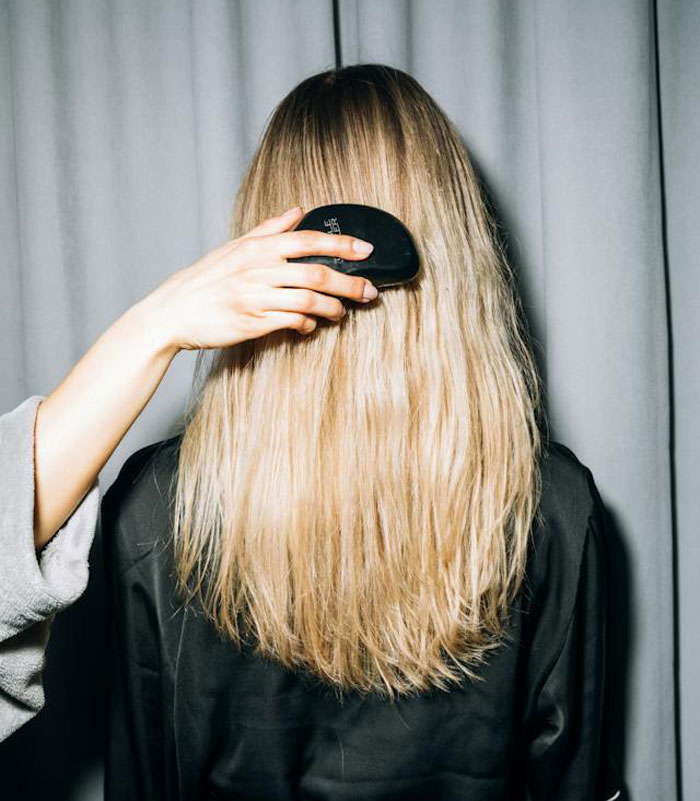 Woman helping detangle daughter's friend's blonde hair with a black brush against a gray curtain background.