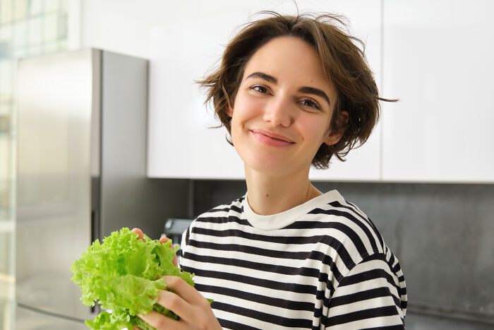 Smiling vegan teen holding fresh lettuce in a modern kitchen, embracing her plant-based diet confidently.