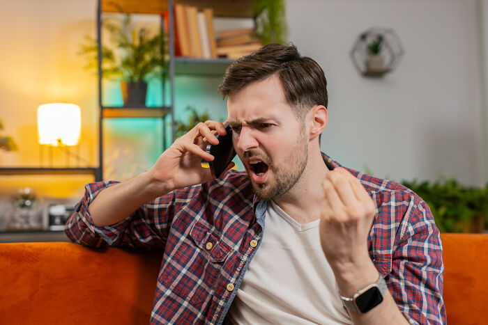 Angry man in plaid shirt talking on phone, expressing frustration in a brightly lit living room setting.