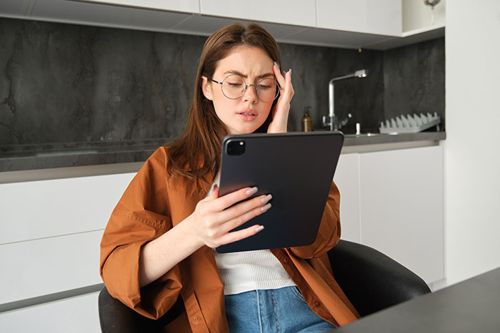 Young woman in glasses reading tablet with a worried expression after finding secret texts about her appearance. Young woman in glasses reading tablet with a worried expression after finding secret texts about her appearance.