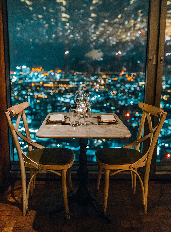Mesa de jantar romântica posta para dois com copos e guardanapos com vista para a paisagem noturna da cidade, refletindo um cenário de namoro idiota.