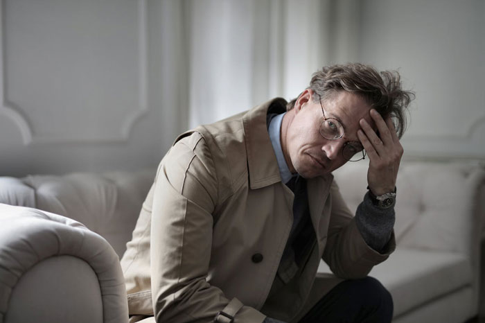 Man in beige trench coat and glasses sitting on couch, looking distressed and unsure about affair partner's baby situation. Man in beige trench coat and glasses sitting on couch, looking distressed and unsure about affair partner's baby situation.