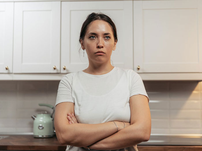 Young woman with crossed arms and a serious expression in a kitchen, reflecting uncertainty about family relationships. Young woman with crossed arms and a serious expression in a kitchen, reflecting uncertainty about family relationships.