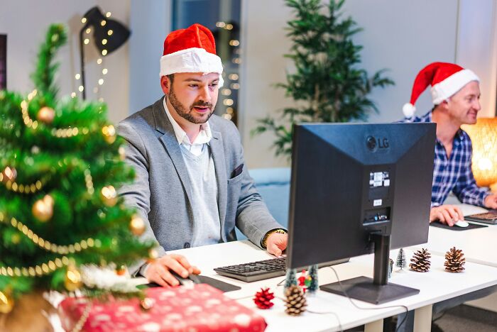 Man wearing Santa hat working at computer in decorated office during holiday season, while woman plans outing with husband work