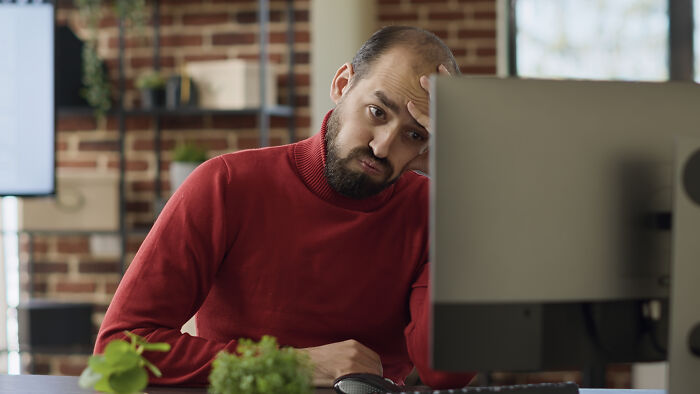 Man working at computer looking stressed while woman plans outing in the background of home office setting Man working at computer looking stressed while woman plans outing in the background of home office setting