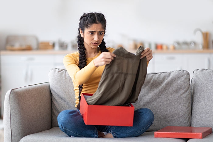 Young woman confused about Christmas gifts from boyfriend holding missing items inside red gift box on sofa.