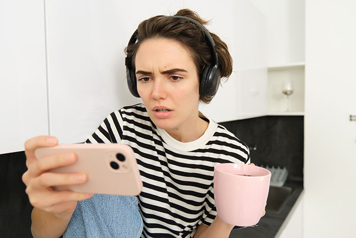 Young woman with headphones holds phone and pink mug, looking shocked while reading social media about coworker conflict.