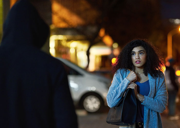 Woman standing on a dimly lit street looking concerned as a man in a hoodie approaches, highlighting stalker fear period pad. Woman standing on a dimly lit street looking concerned as a man in a hoodie approaches, highlighting stalker fear period pad.