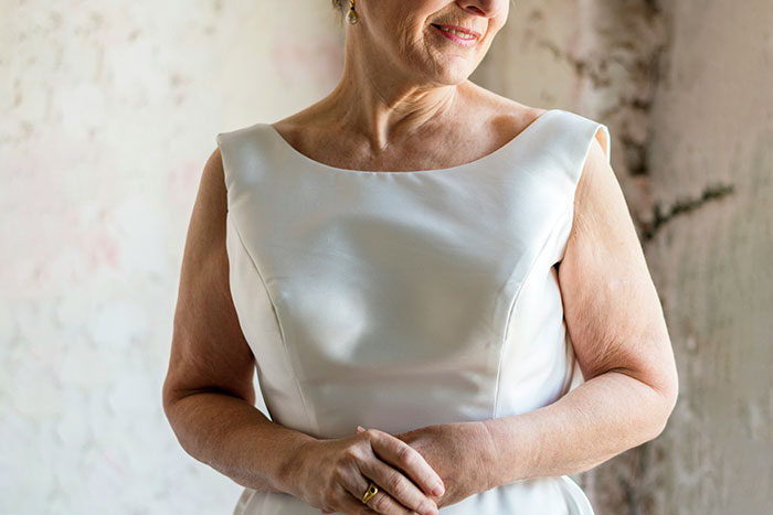 Middle-aged woman wearing a white dress, preparing for a son's wedding, looking thoughtful and slightly smiling.