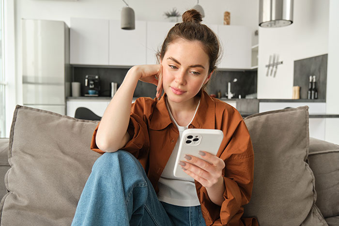 Woman in casual outfit sitting on couch looking thoughtfully at smartphone, reflecting on wedding photo controversy. Woman in casual outfit sitting on couch looking thoughtfully at smartphone, reflecting on wedding photo controversy.