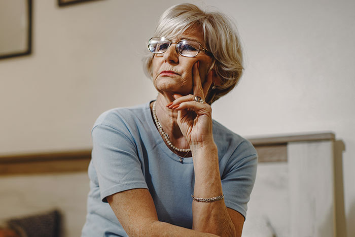 Older woman with glasses thoughtfully resting her face on her hand, reflecting on wedding dress controversy with bride and son. Older woman with glasses thoughtfully resting her face on her hand, reflecting on wedding dress controversy with bride and son.