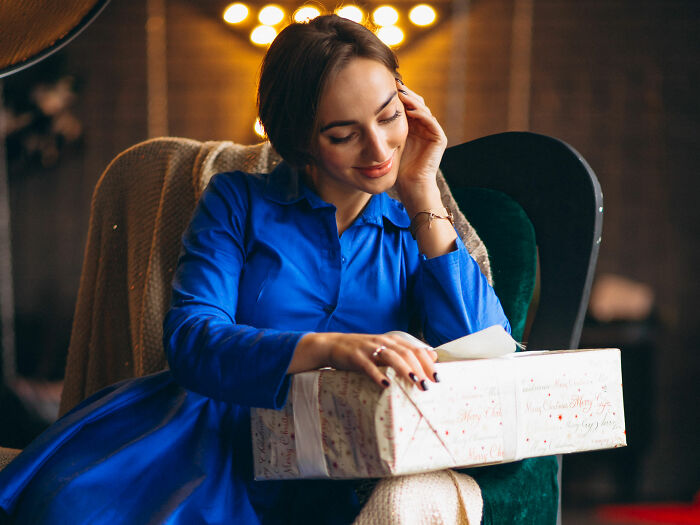 Woman in blue dress sitting in cozy armchair, holding holiday gift that might reveal painful truth about relationship. Woman in blue dress sitting in cozy armchair, holding holiday gift that might reveal painful truth about relationship.