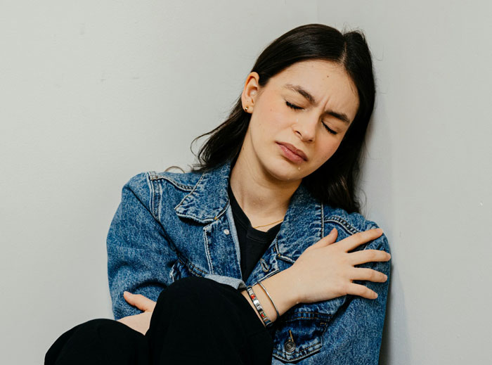 Woman in denim jacket sitting against a wall looking upset, illustrating a woman blinded by flowers from her son. Woman in denim jacket sitting against a wall looking upset, illustrating a woman blinded by flowers from her son.