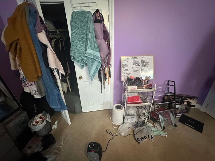 Cluttered bedroom with pink and purple tones, clothes hanging on door, and various items scattered on floor and shelves.